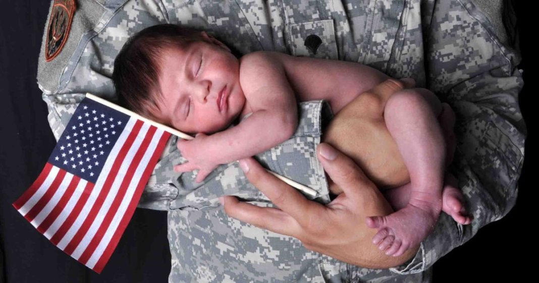 This image shows an American soldier holding a newborn baby who is holding onto an American flag while sleeping.